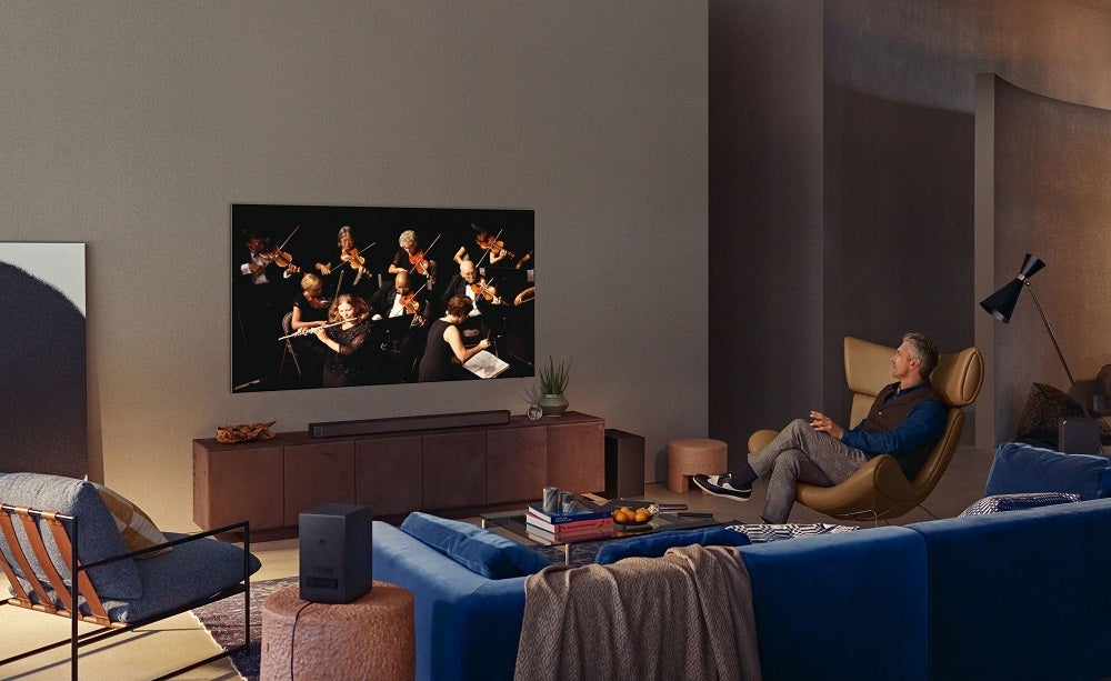 Un televisor Samsung Neo QLED 4K negro colgado de una pared con un anciano sentado en una silla y viendo la televisión