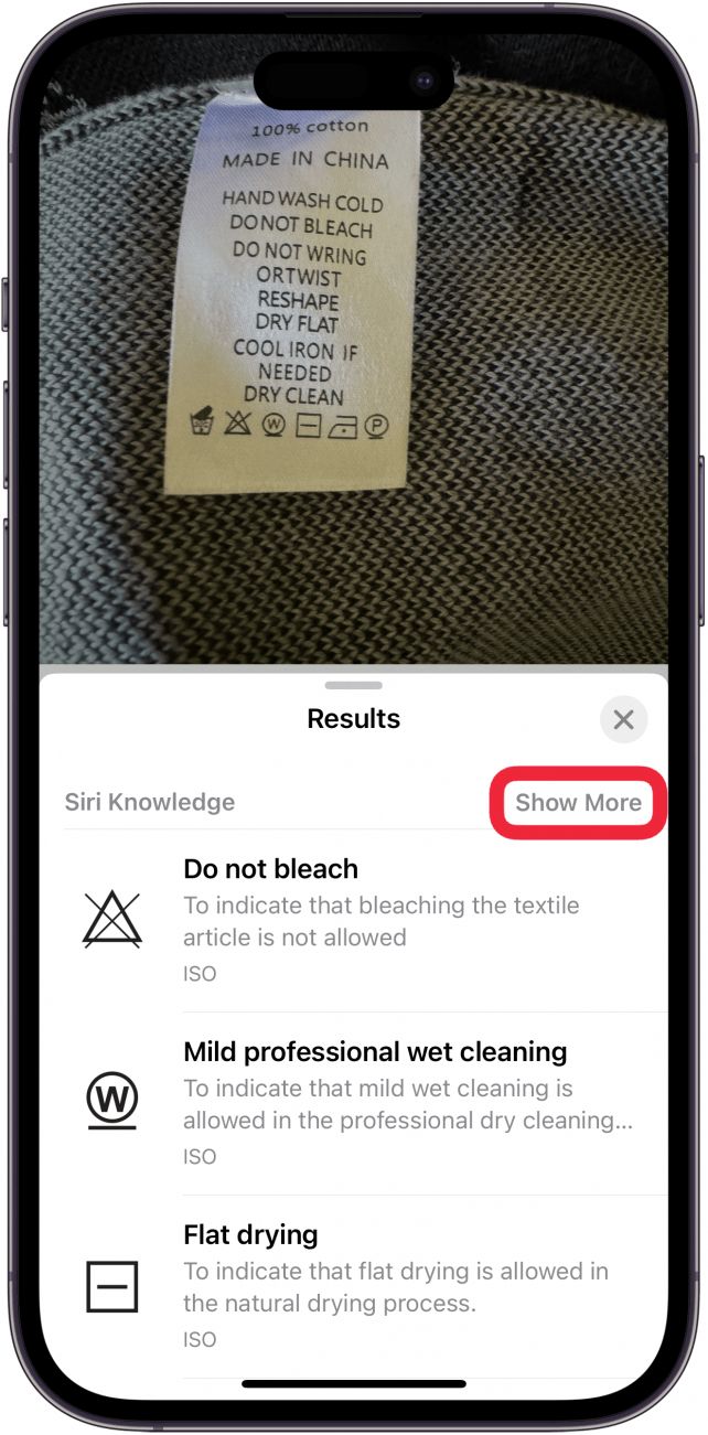 Aplicación de fotos para iPhone que muestra una foto de una etiqueta de lavandería en un suéter, con el menú de búsqueda visual abierto, que muestra diferentes símbolos de lavandería y su significado.  hay un cuadro rojo alrededor del botón mostrar más